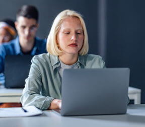 A young woman with blonde hair focusing on a laptop at a desk in a classroom setting.