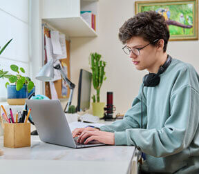 A person wearing glasses and a light-colored sweatshirt sits at a desk using a laptop. They have headphones around their neck, and the desk features various items including plants, a globe, and a microphone.