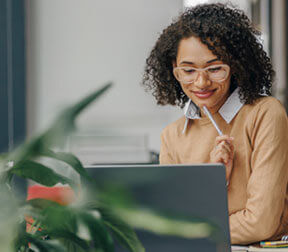 A person with curly dark hair and glasses smiles while looking at a laptop, holding a pen to their chin with a green plant in the blurred foreground.