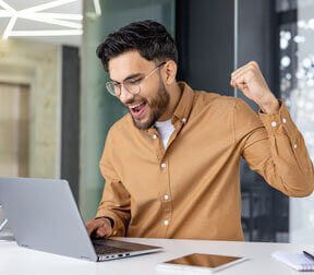 A person with dark hair and a beard cheers with a fist pump while looking at a laptop. They wear glasses and a tan shirt, with a tablet and notebook on the white desk in a bright office.