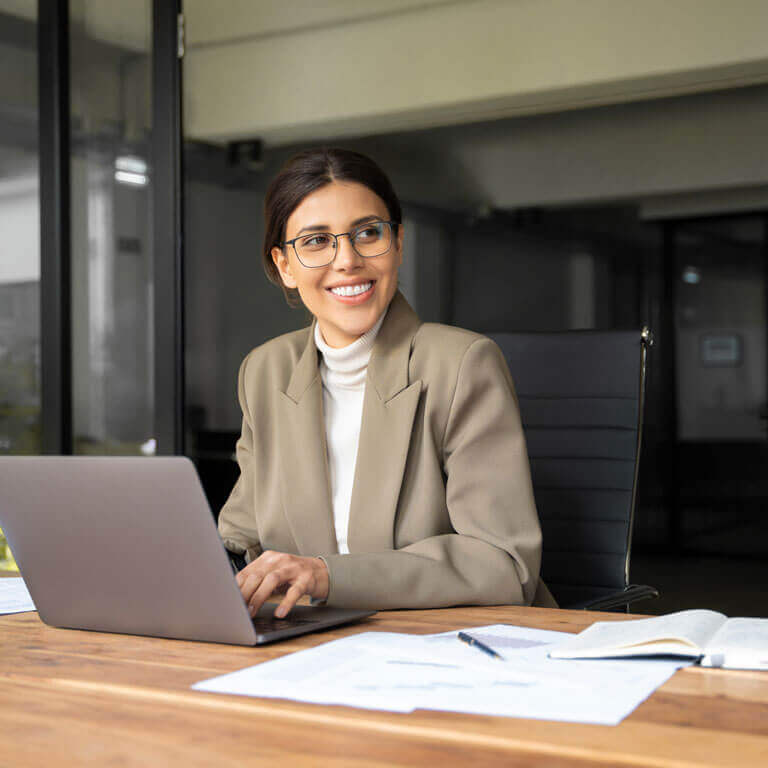 A person with glasses and a tan blazer smiles while looking away from their laptop. They sit at a wooden desk with papers and a notebook in a modern office.