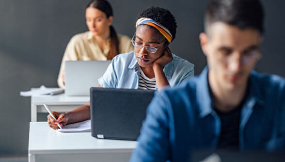 A person with glasses and a headband writes in a notebook next to a laptop. Other individuals are visible working at desks in a classroom setting.